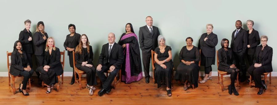 The Van Zyl Retief Incorporated team in black, seated and standing on wooden chairs and floorboards against a sage-green wall, Port Shepstone.