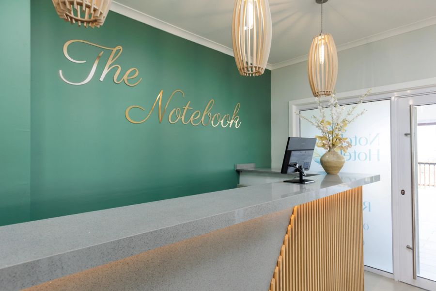 Hotel reception desk with 'The Notebook' signage in gold script on a green wall, pendant lights, and a floral arrangement on the counter.
