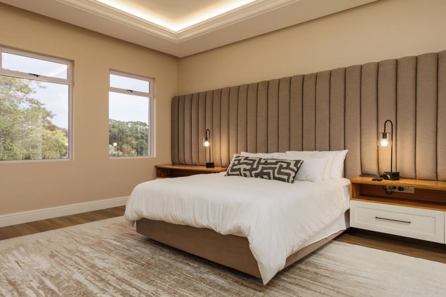Bedroom in a modern home with a channel-tufted headboard, soft lighting, and large windows looking onto trees.