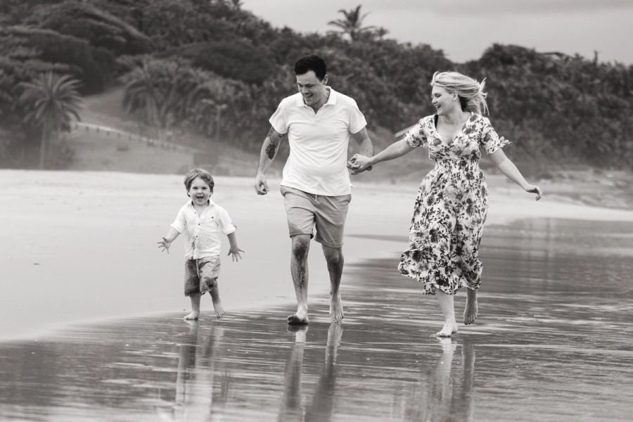 Black and white photo of a family of three laughing and holding hands while walking along the beach.