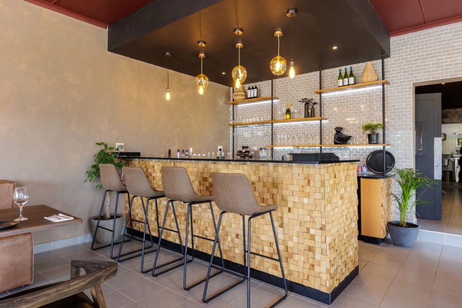 Bar at Feje’s Restaurant in Umtentweni, with a timber-block counter, tall leather bar stools, pendant lights and glass shelving against a white brick wall.
