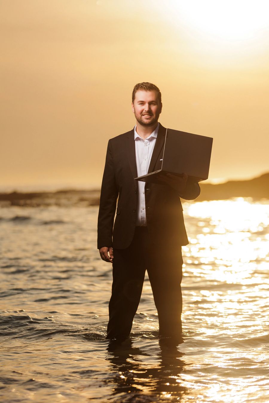 Businessman in a suit standing knee-deep in the ocean at sunset, holding an open laptop under his arm.