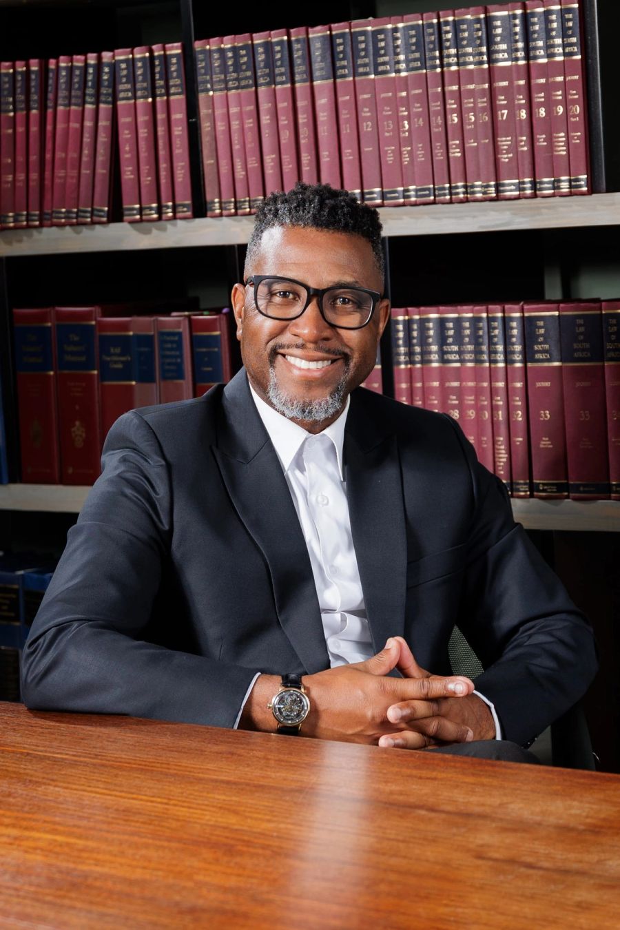 Corporate portrait of a man in a dark suit and glasses seated at a wooden table in front of shelves of law books, Shelly Beach.