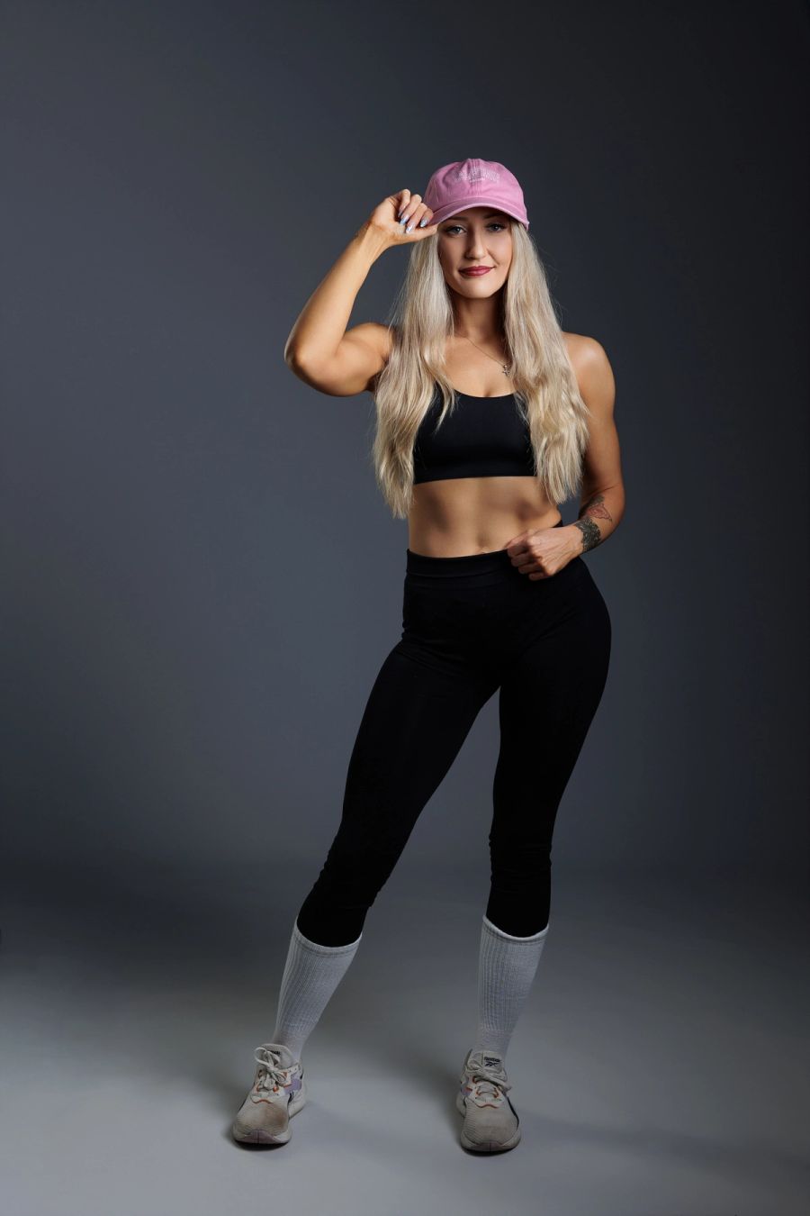 Woman in black activewear and a pink cap posing in a studio against a grey backdrop.