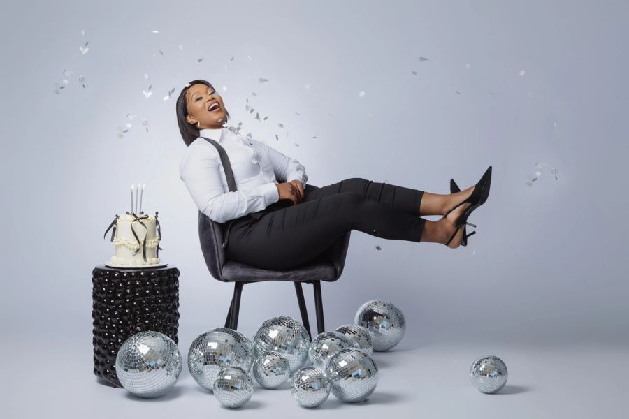 Woman in a white shirt and black trousers laughing while reclining in a chair surrounded by disco balls, confetti, and a birthday cake.