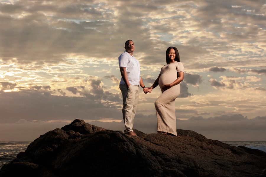 Expecting couple holding hands on a rocky outcrop above the sea under a dramatic sunset sky, Southport.