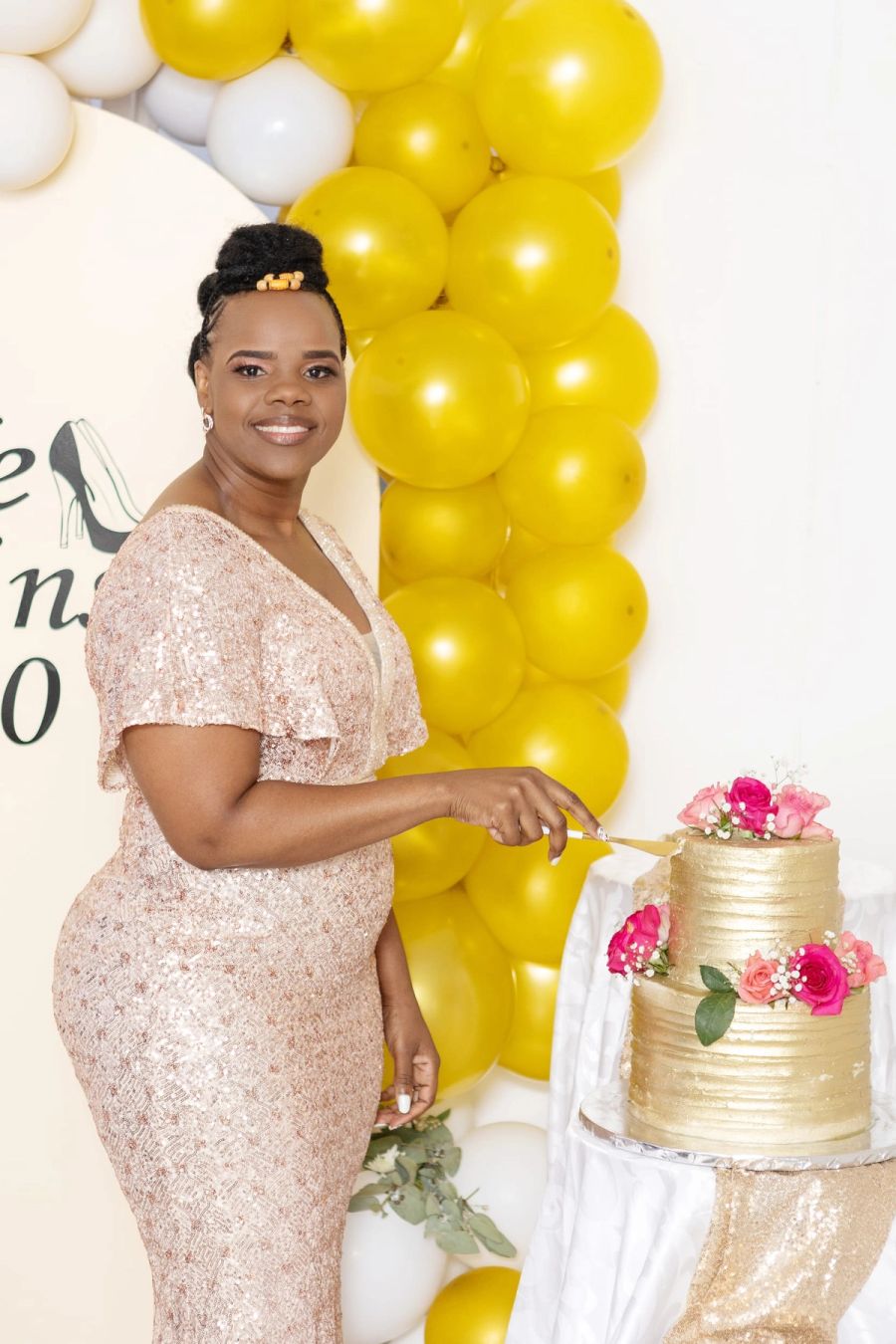 Woman in a sparkling pink dress cutting a gold-tiered birthday cake beside a yellow and white balloon arch, Port Edward.