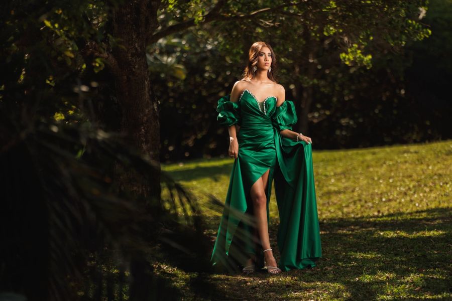 Teenage girl in an emerald green gown standing in dappled light beneath trees.
