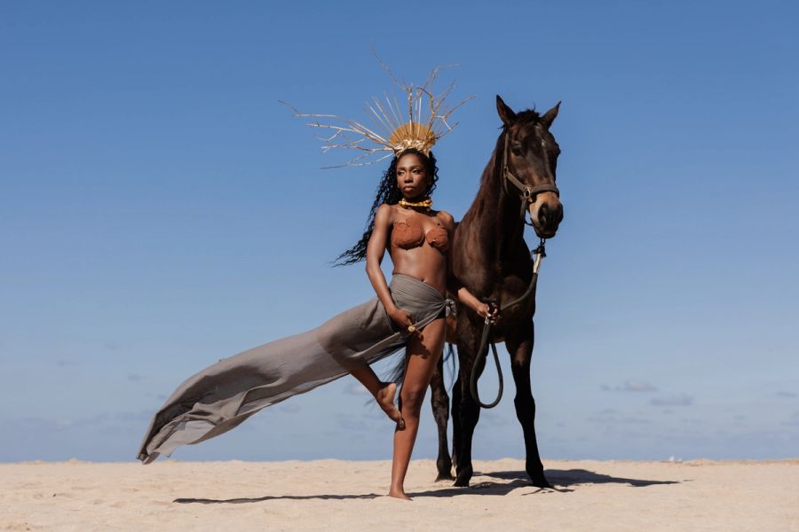 Woman in a sculptural headdress and flowing fabric standing beside a horse on the beach against a clear blue sky.
