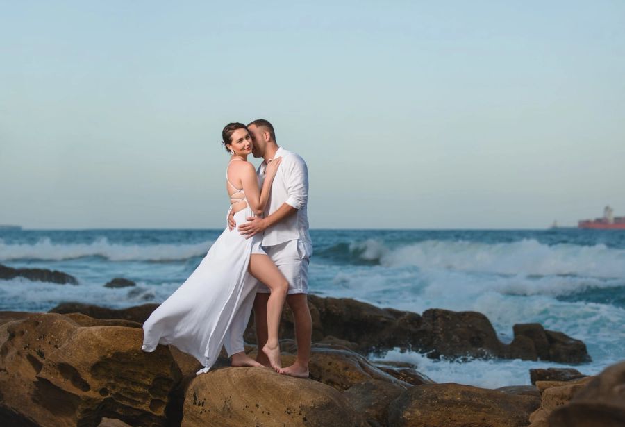 Couple embracing on the rocks at the water’s edge, the woman in a long white dress, with waves breaking behind them, Umhlanga.