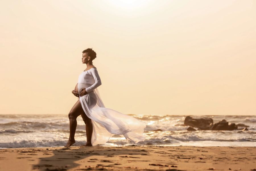 Expectant mother in a flowing white sheer gown standing on the beach at sunrise, the sea behind her, Port Edward.