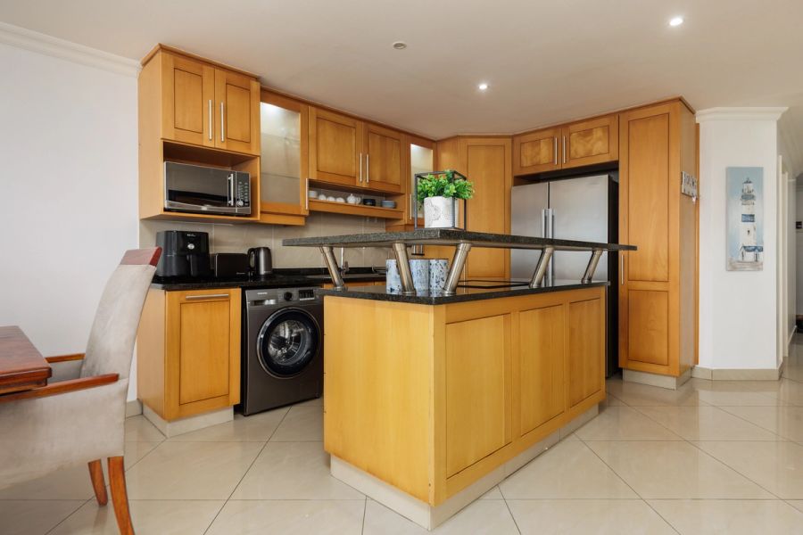 Modern kitchen with honey-toned wood cabinetry, a central island, stainless steel appliances, and beige tile flooring.