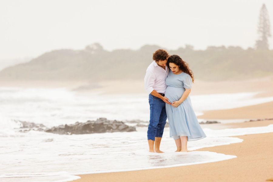Couple standing in the surf embracing, the man's hands resting on his pregnant partner's belly.