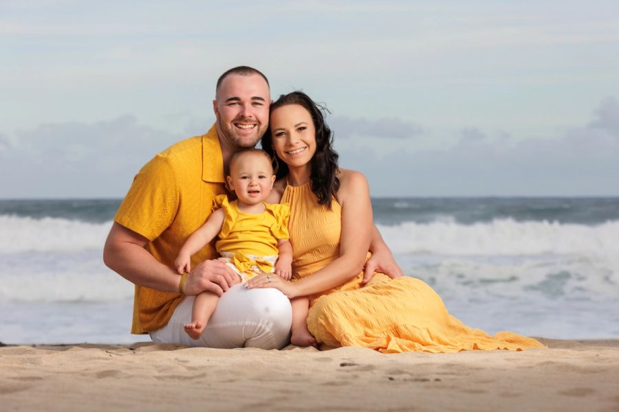 Family of three in matching yellow outfits sitting together on the beach with ocean waves in the background.