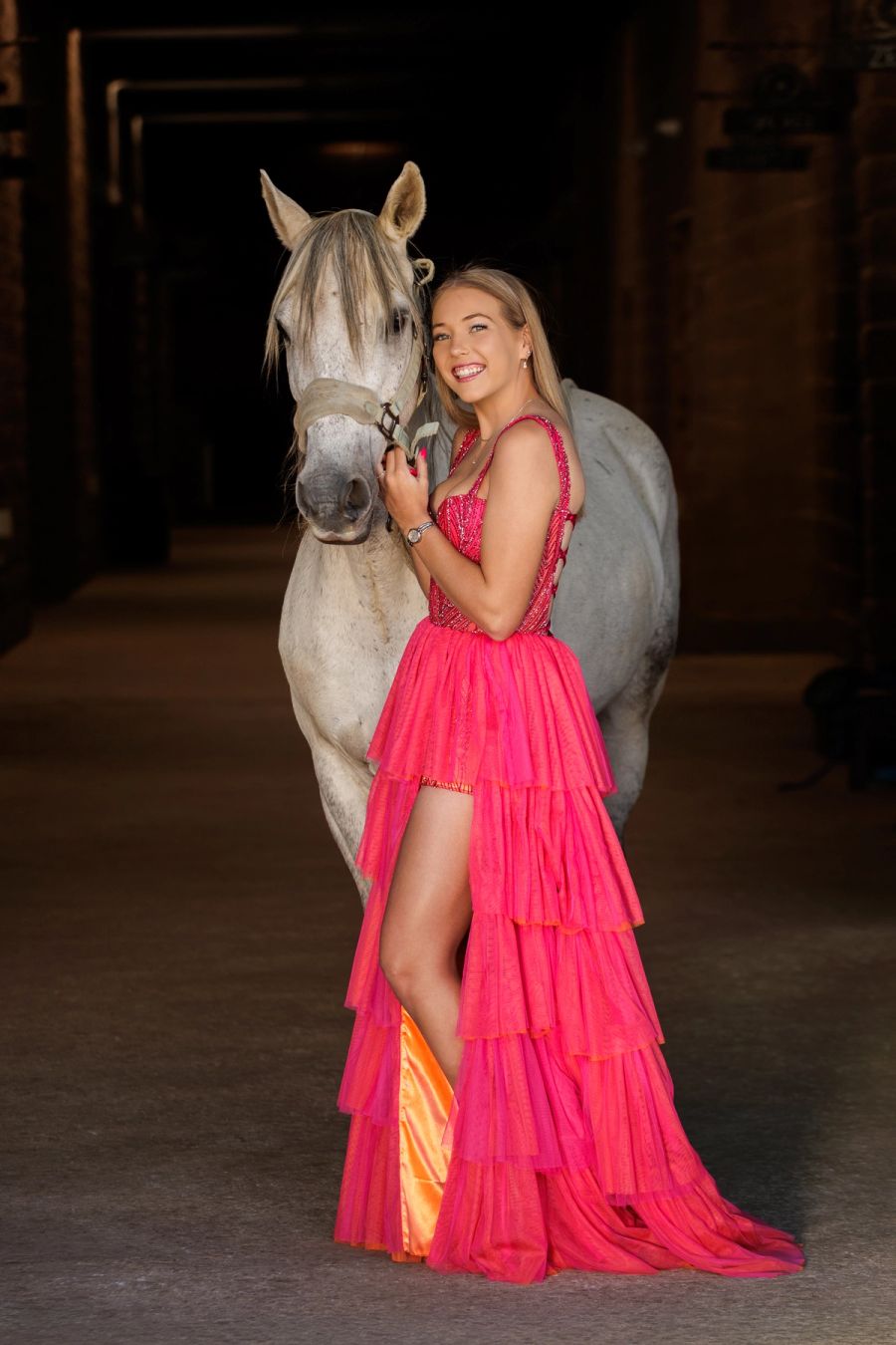 Teenage girl in a bright pink ruffled gown smiling beside a white horse in a stable doorway.