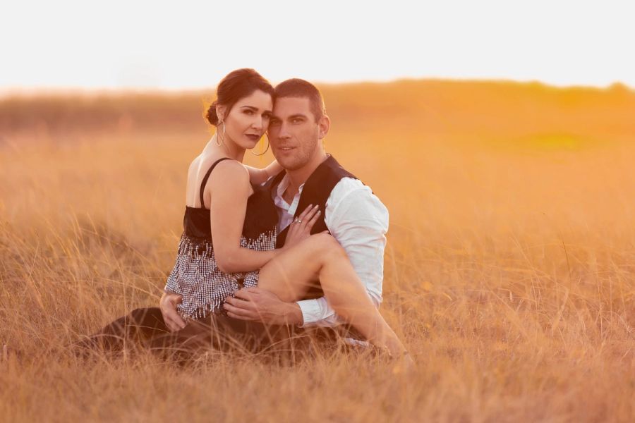 Couple sitting close together in golden grass at sunset, smiling at the camera, Margate.
