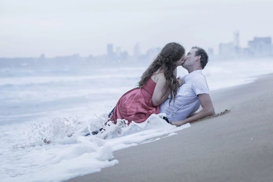 Couple kissing as a wave washes over them on the beach, with a hazy city skyline in the distance, Durban.