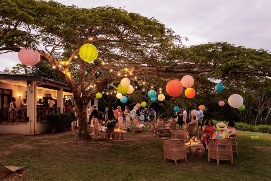 Guests at an outdoor year-end function under a large tree hung with colourful paper lanterns and string lights, Oslo Beach.