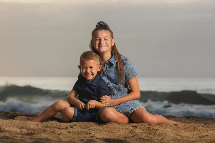 Smiling brother and sister sitting together on the sand with waves breaking behind them, Leisure Bay.