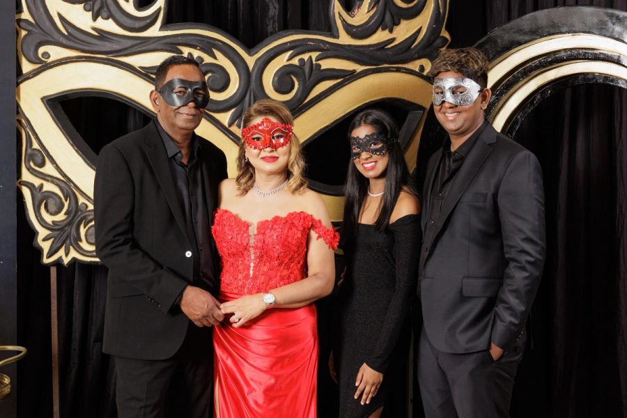 Four guests in black and red evening wear and masquerade masks posing in front of a gold mask backdrop, Oslo Beach.