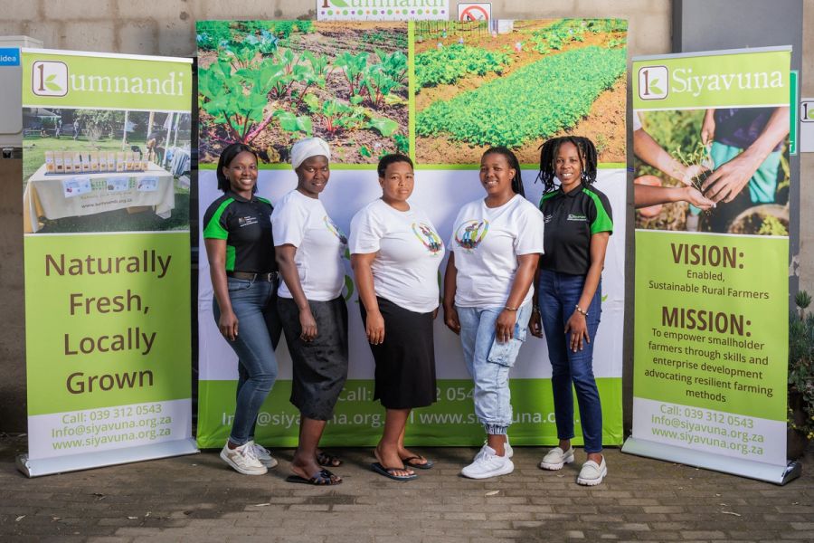 Five team members in branded T-shirts standing together in front of a Kumnandi and Siyavuna display banner outdoors.