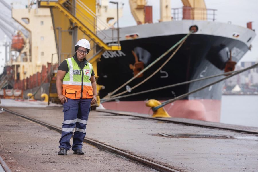 Female manager in a hard hat and hi-vis vest standing on the quayside in front of a moored cargo ship at the Port of Durban.