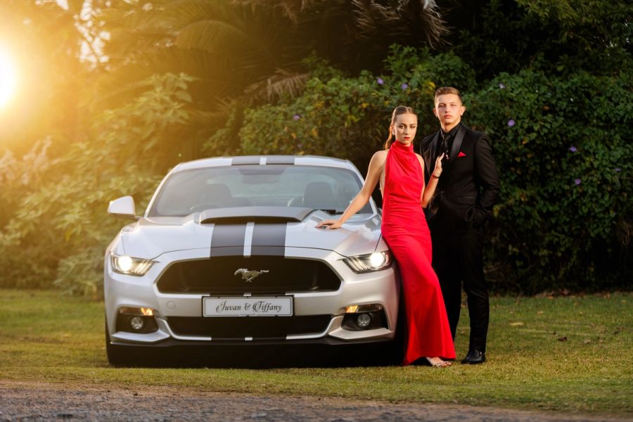Matric dance couple in red and black formalwear posing beside a silver muscle car at sunset.