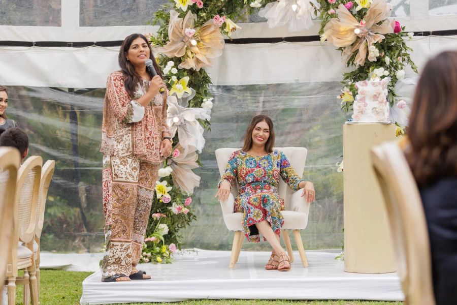 Guest speaking into a microphone beside the guest of honour seated in a floral-arched chair under a garden marquee at a 60th birthday celebration, Oslo Beach.