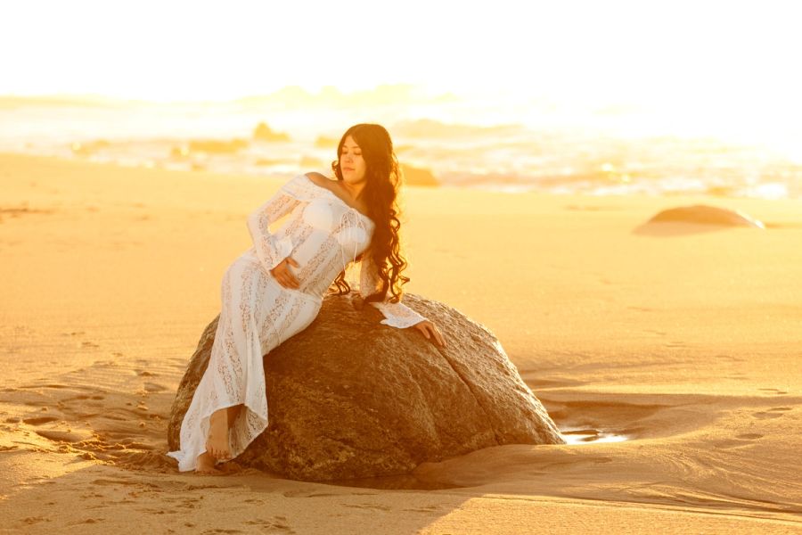 Pregnant woman in a white lace gown leaning against a large beach rock at golden hour.