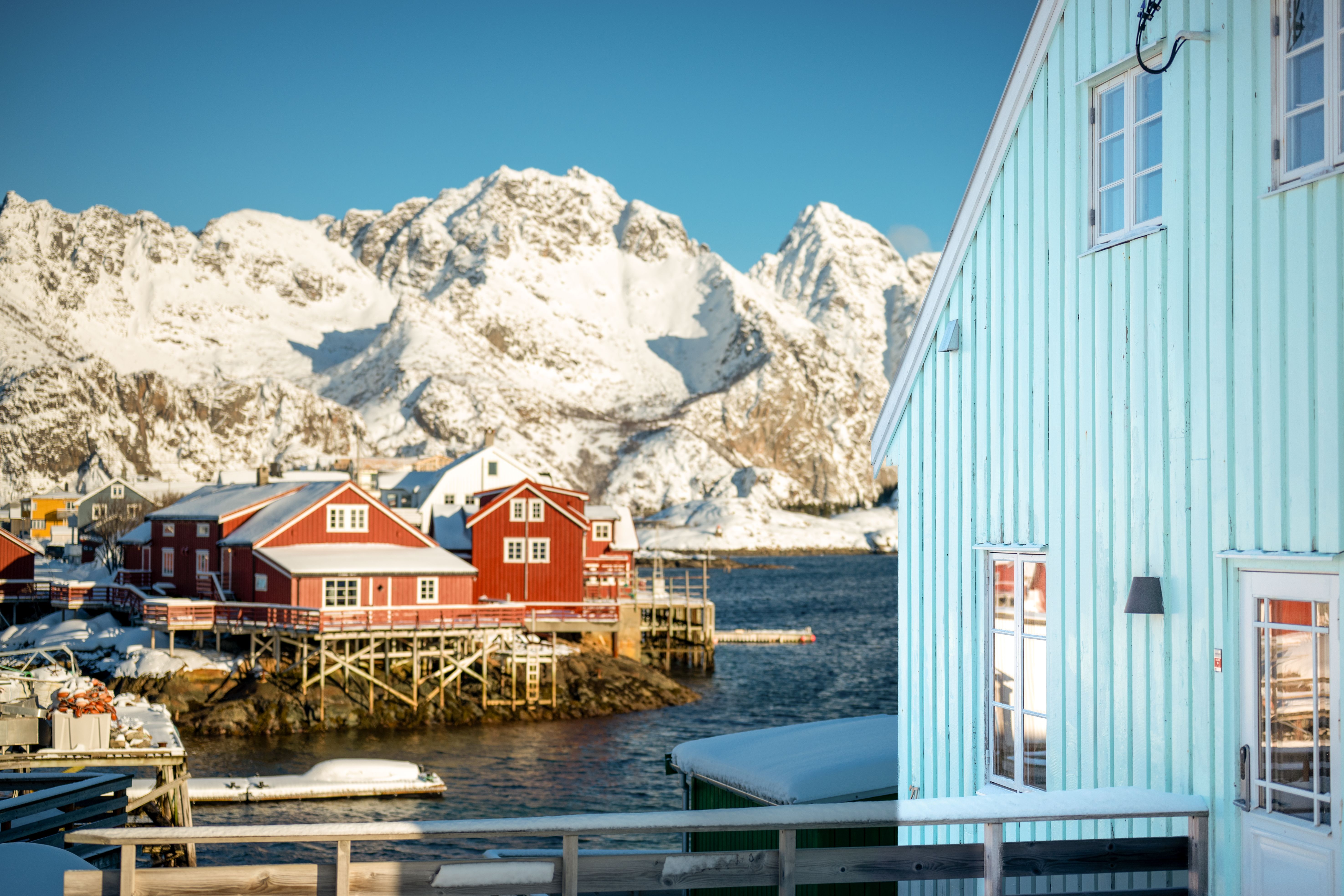 Arctic Coliving - a turquoise lodge on the water in Henningsvær, Norway with mountains in the background.
