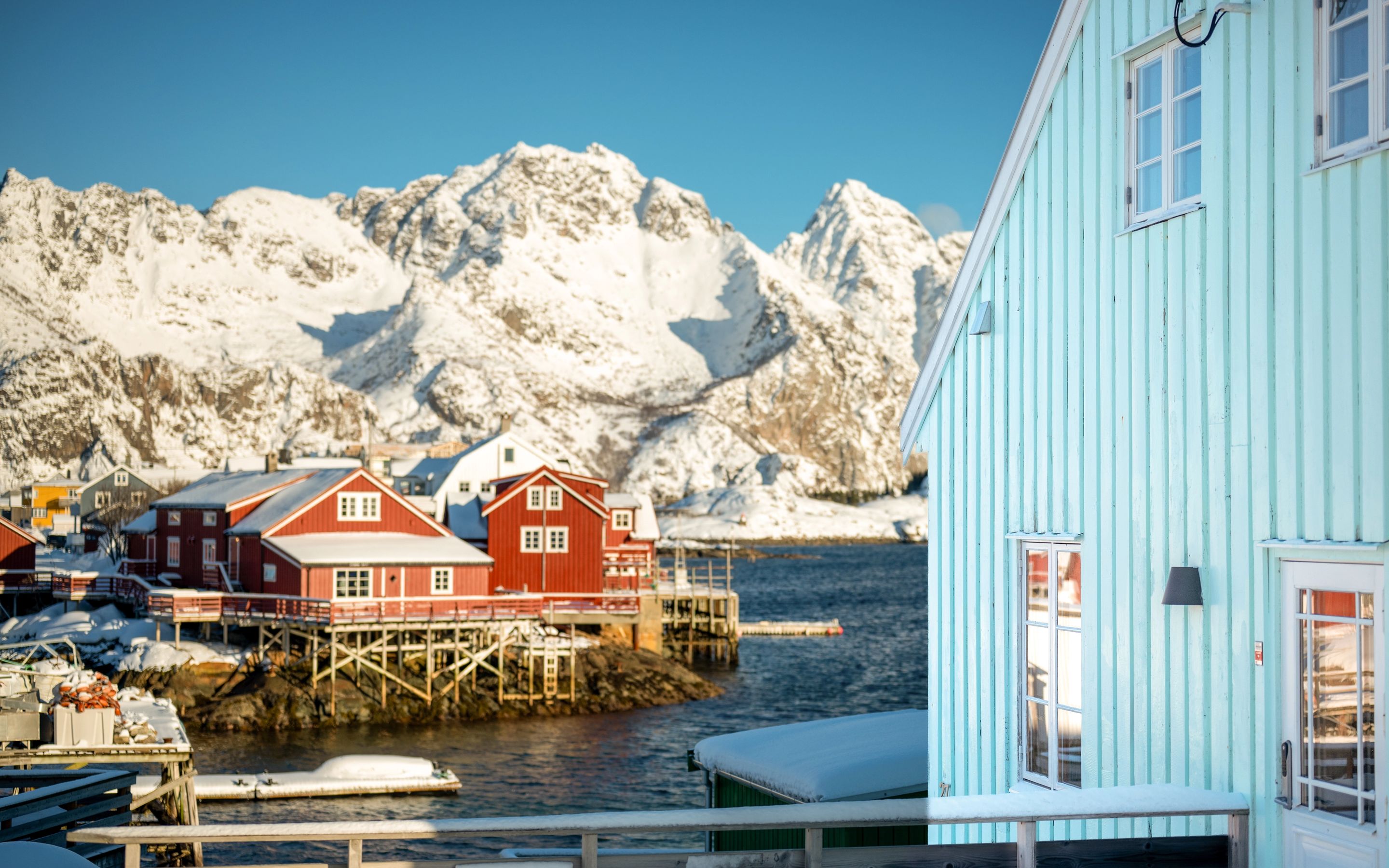 Arctic Coliving - a turquoise lodge on the water in Henningsvær, Norway with mountains in the background.