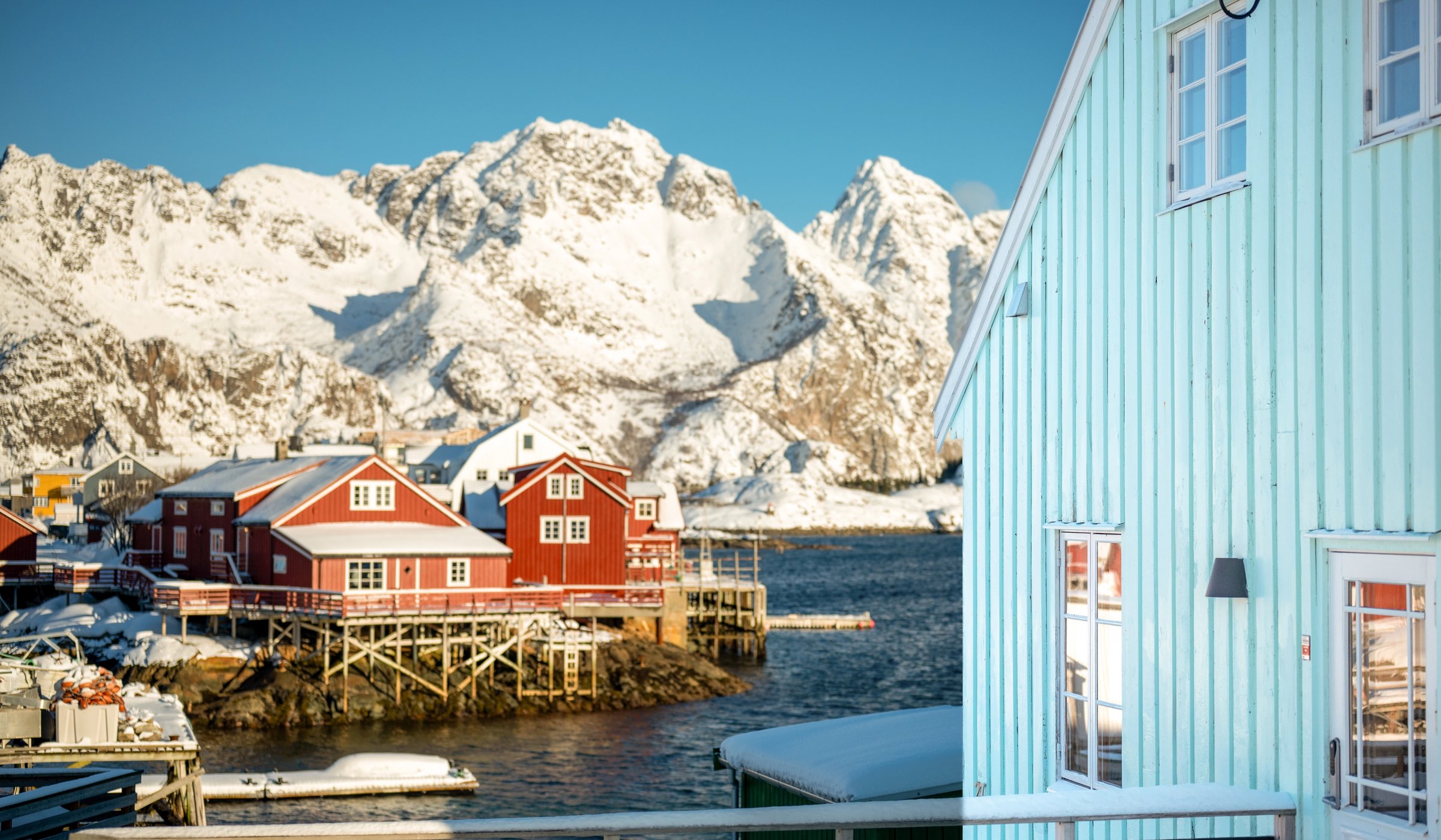 Arctic Coliving - a turquoise lodge on the water in Henningsvær, Norway with mountains in the background.