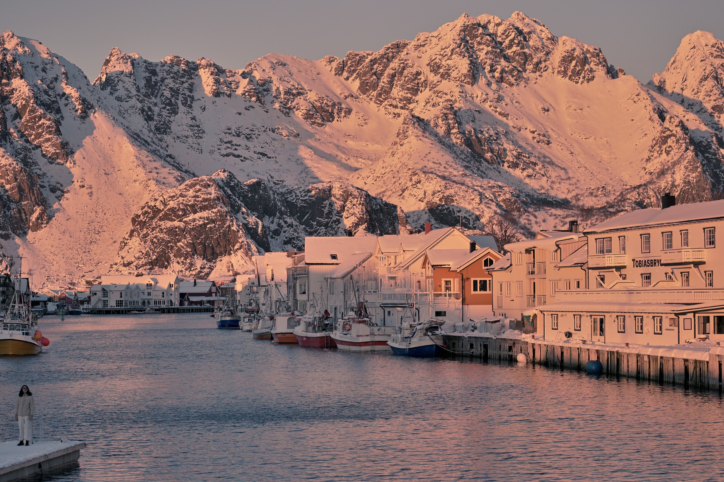 Henningsvær harbour in Lofoten at sunrise, with snow-covered mountains behind.