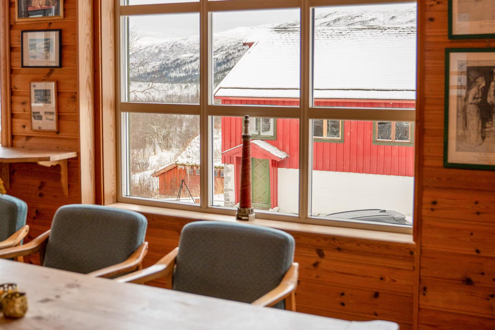Window view from a wooden room of a snowy landscape with red houses, distant mountains, and a decorative red object on the sill.