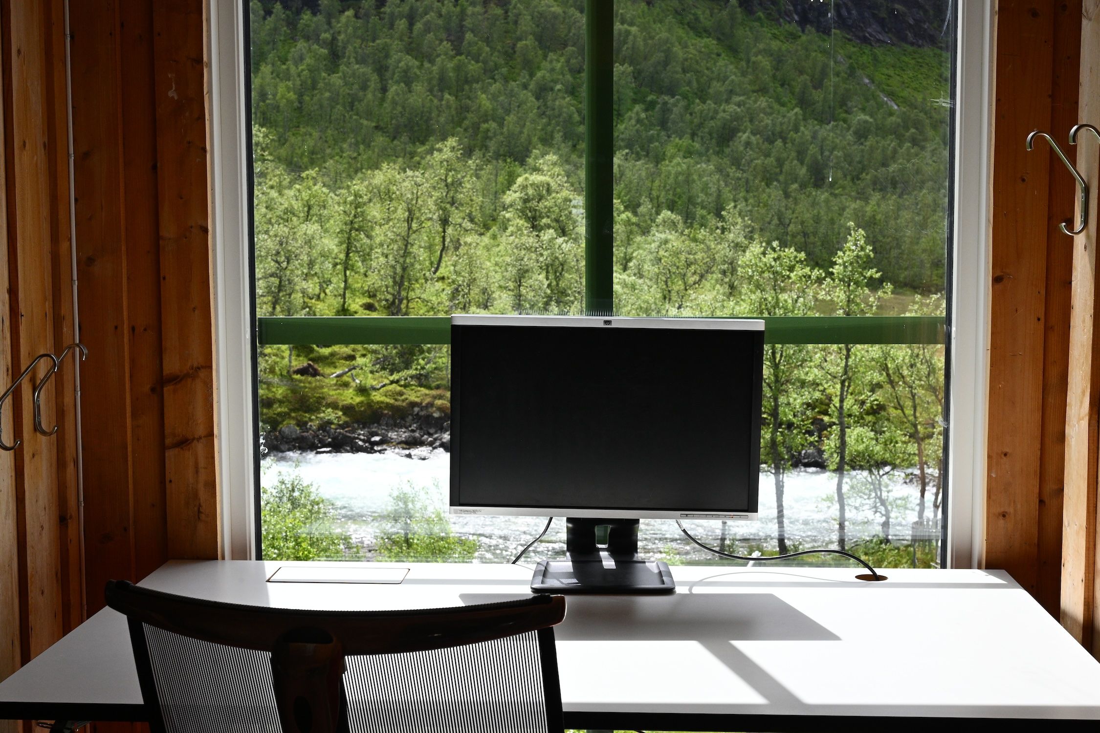 A desk with a monitor and chair by a window overlooking a river and a green forest.