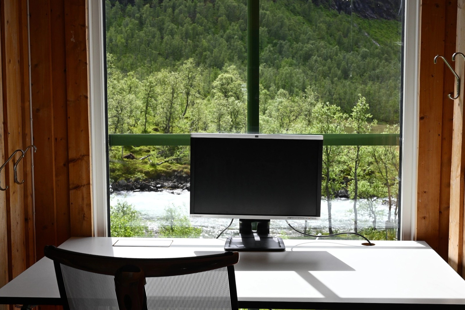A desk with a monitor and chair by a window overlooking a river and a green forest.
