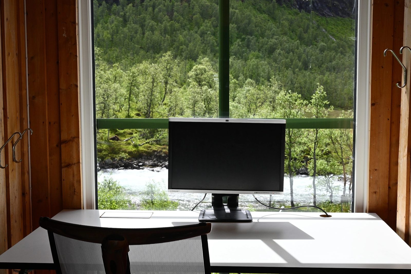 A desk with a monitor and chair by a window overlooking a river and a green forest.