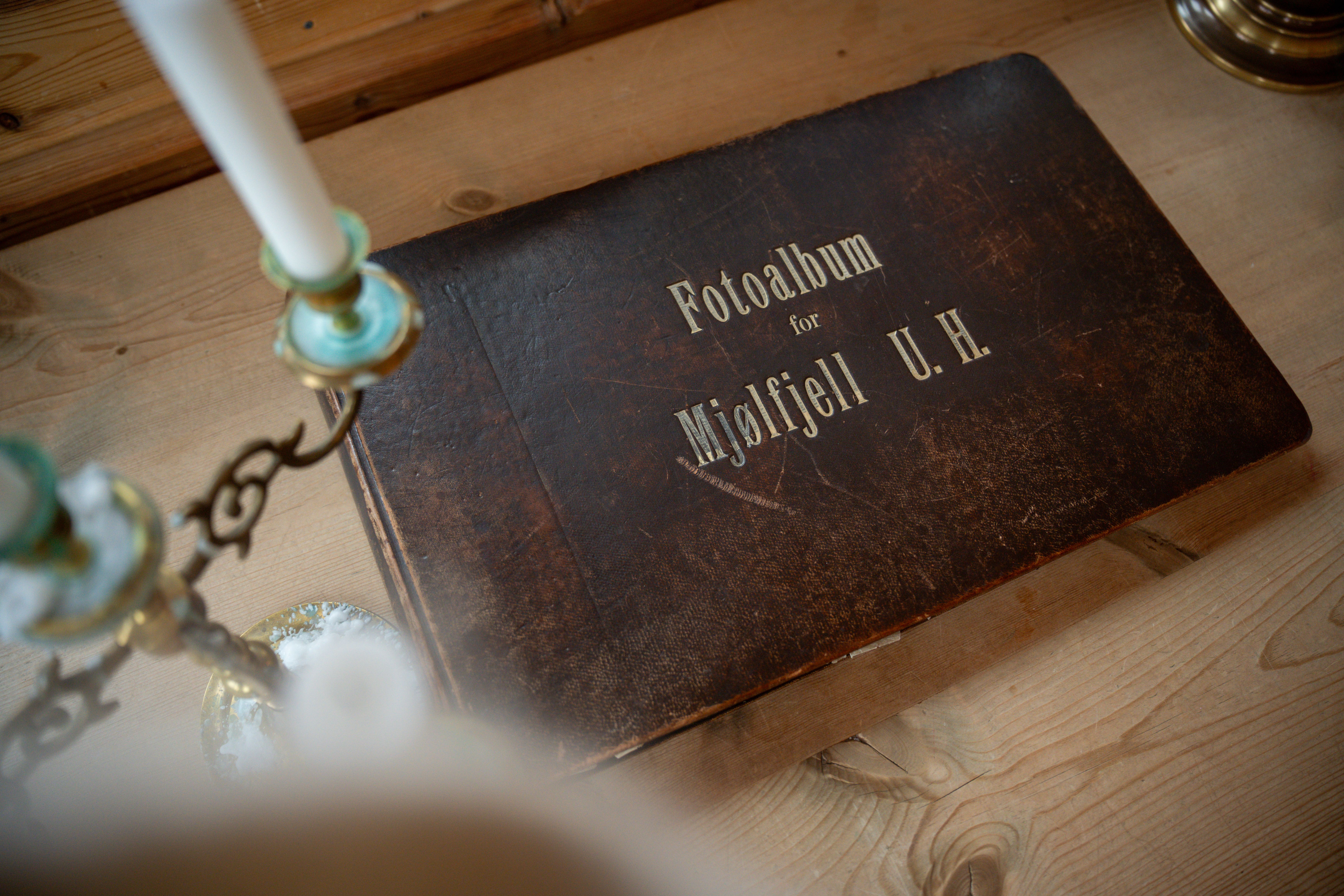 Vintage brown photo album labeled "Fotoalbum for Mjølfjell U. H." on a wooden table with a candelabra.