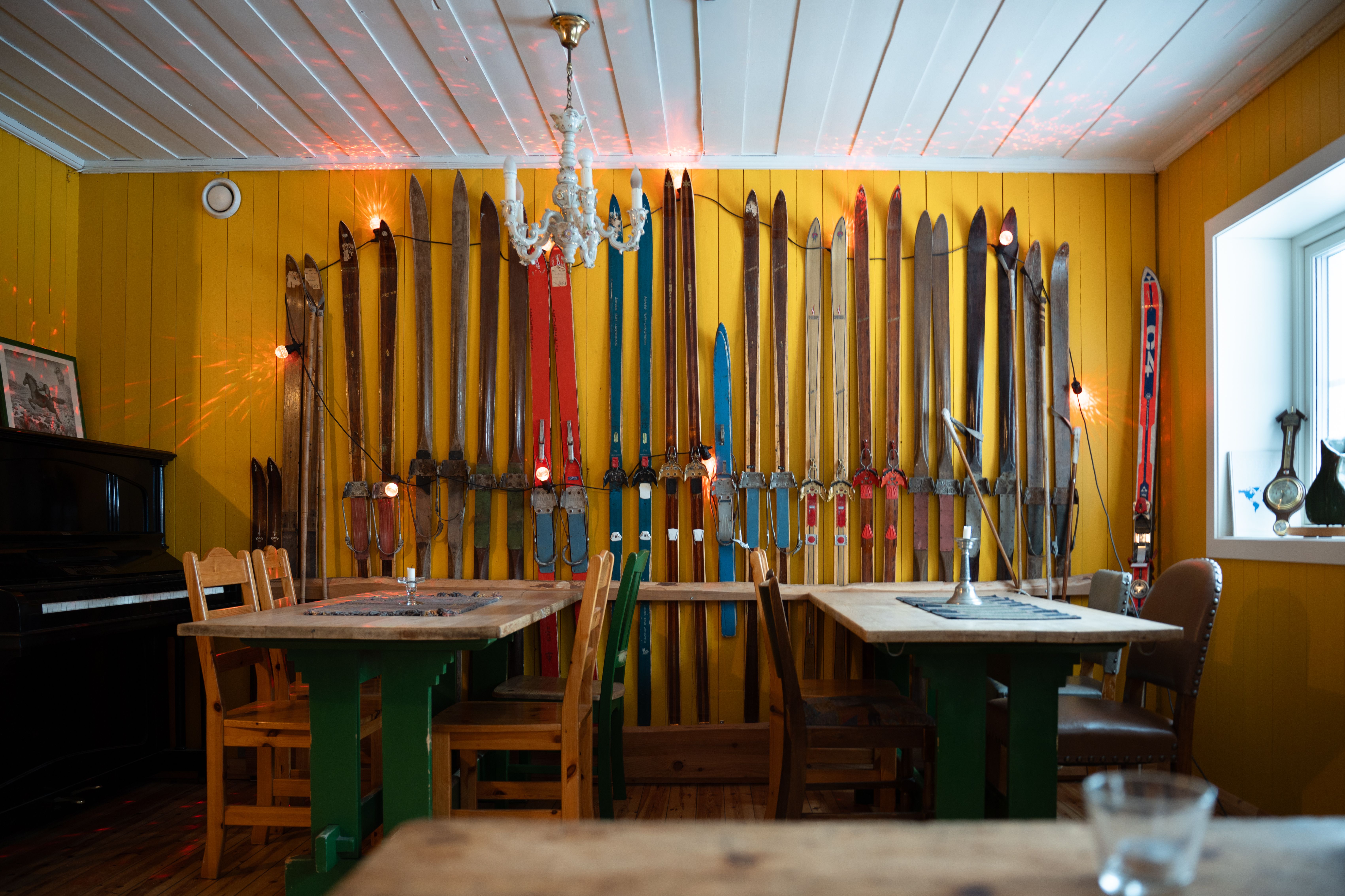 An indoor dining area with bright yellow walls decorated with rows of vintage skis, wooden tables and chairs, a black piano, and a white chandelier.