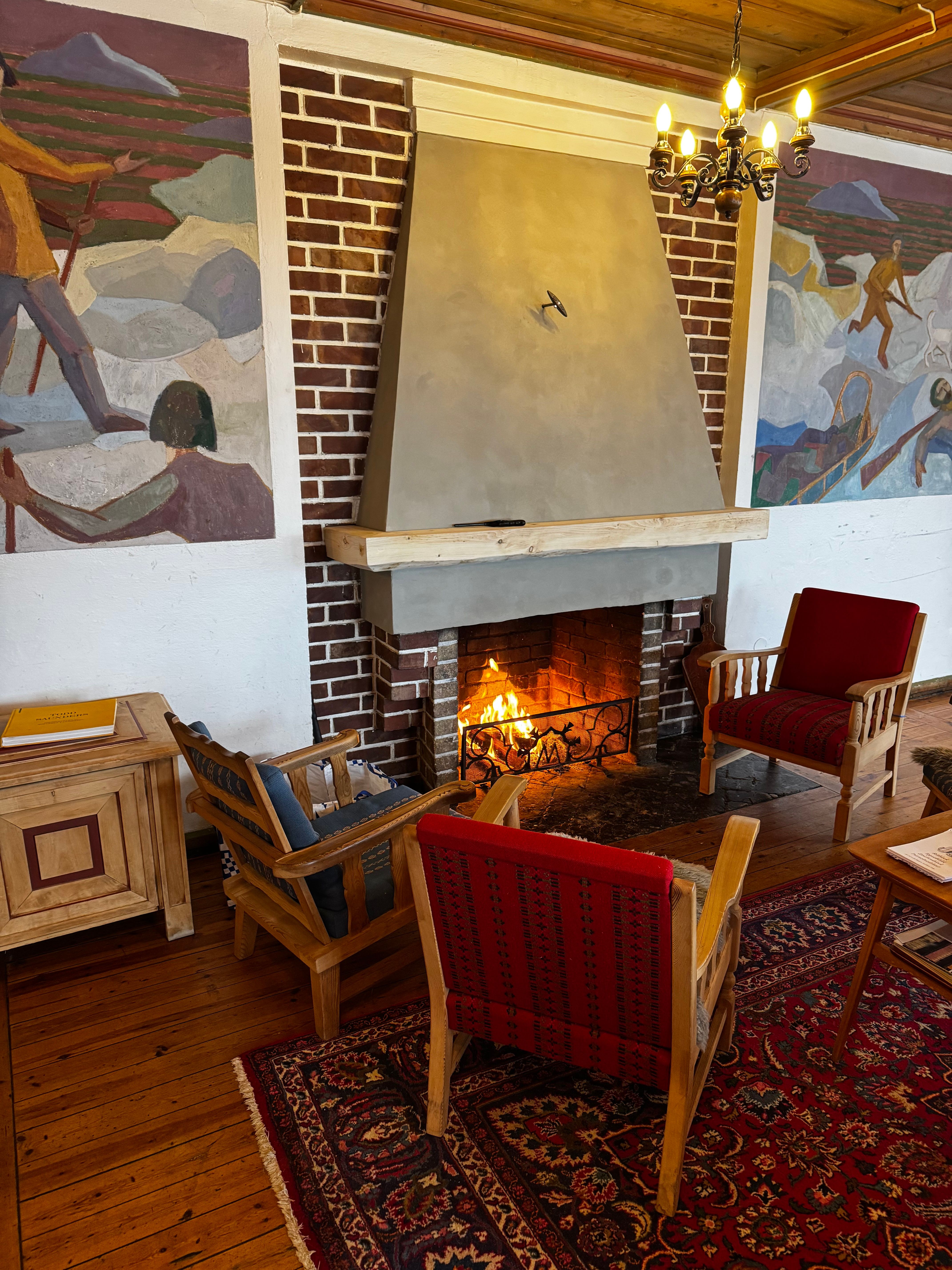 A cozy room with a lit brick fireplace flanked by two large murals, featuring three armchairs, a patterned rug, and a wooden floor.