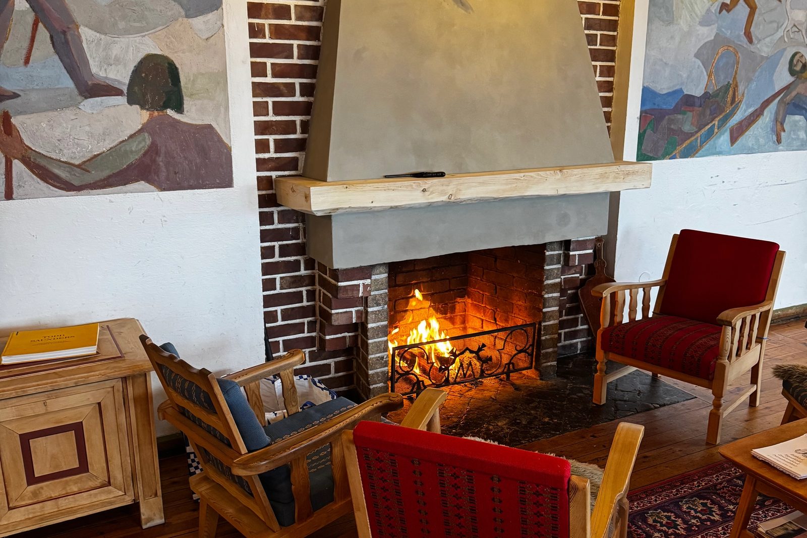A cozy room with a lit brick fireplace flanked by two large murals, featuring three armchairs, a patterned rug, and a wooden floor.