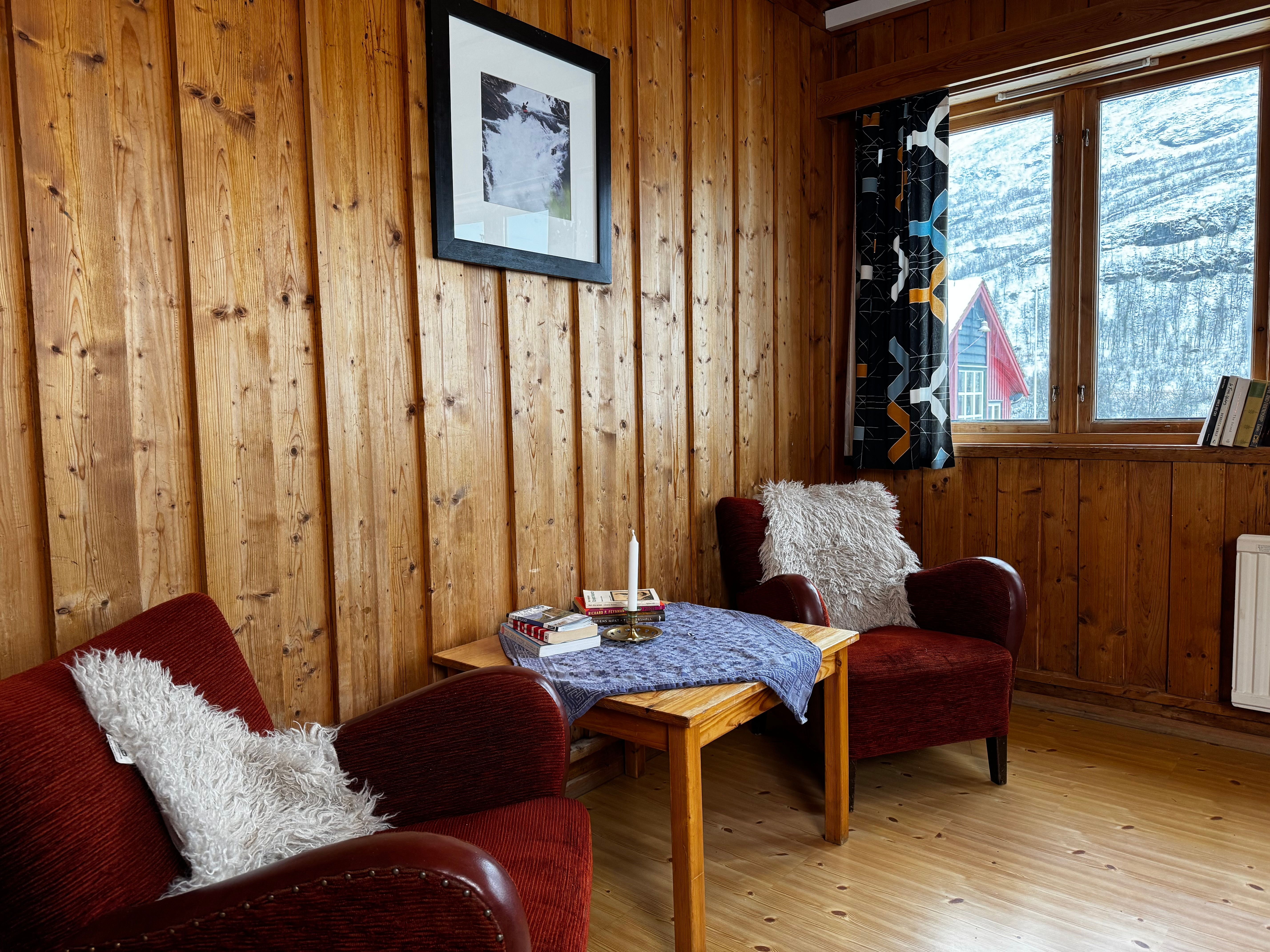 A room with light wood-paneled walls and floor, two red armchairs with white fluffy throws, a wooden side table, and a window overlooking a snowy landscape with a red-roofed building.