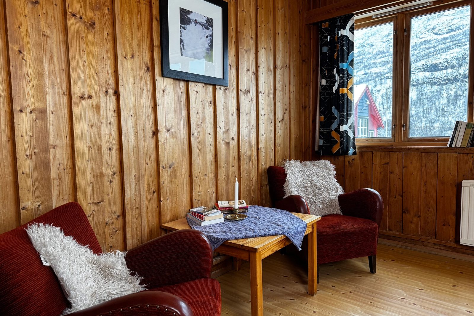 A room with light wood-paneled walls and floor, two red armchairs with white fluffy throws, a wooden side table, and a window overlooking a snowy landscape with a red-roofed building.