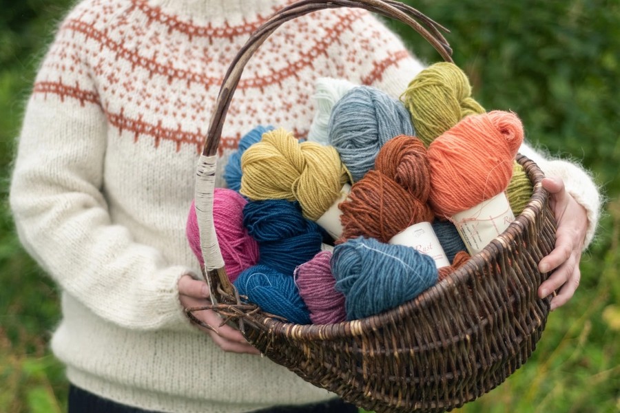 A woman in a traditional cream and orange Norwegian knit carrying a basket of colorful hand-dyed yarn