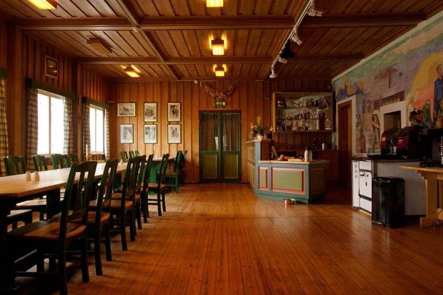 A rustic wooden dining hall with long tables, a bar area, and a colorful mural.