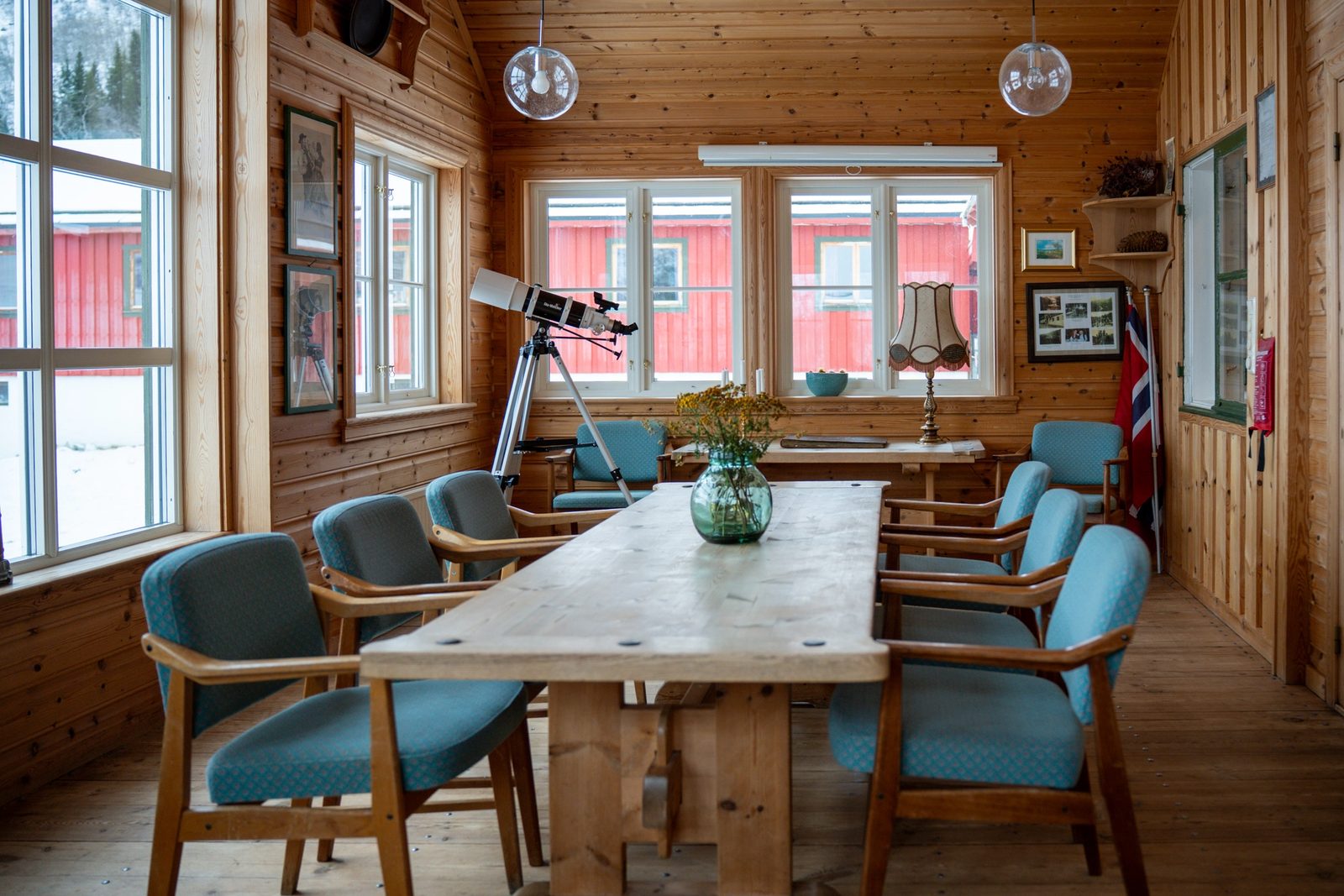 Rustic wood-paneled room with a long dining table, blue chairs, a telescope, and windows showing snowy red cabins.