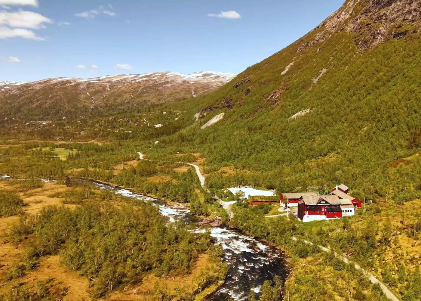 A red-roofed mountain lodge complex by a river in a green valley in Norway, surrounded by forested mountains with distant snowy peaks.