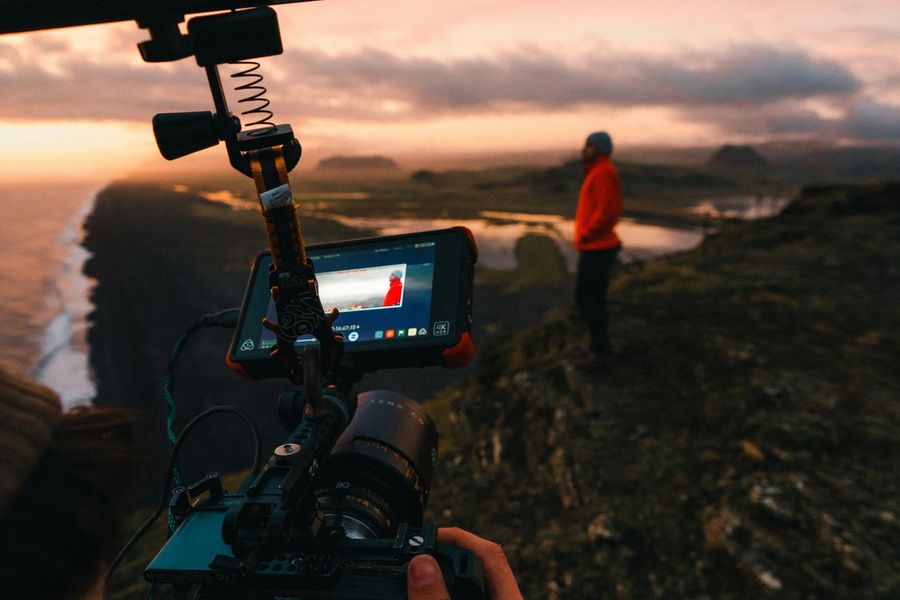 A filmmaker records a person in a red jacket on a rocky coastal cliff at sunset.