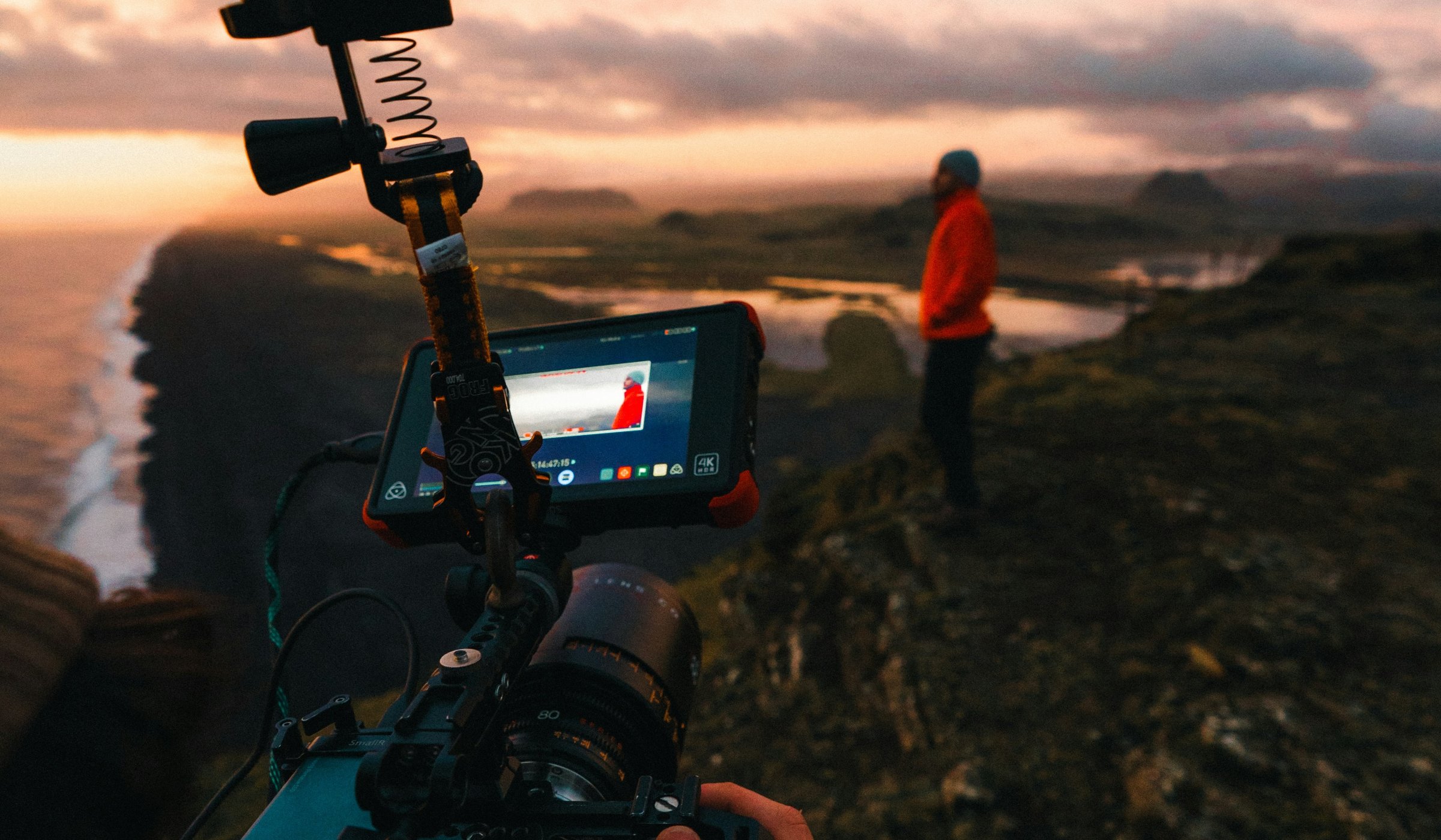 A filmmaker records a person in a red jacket on a rocky coastal cliff at sunset.