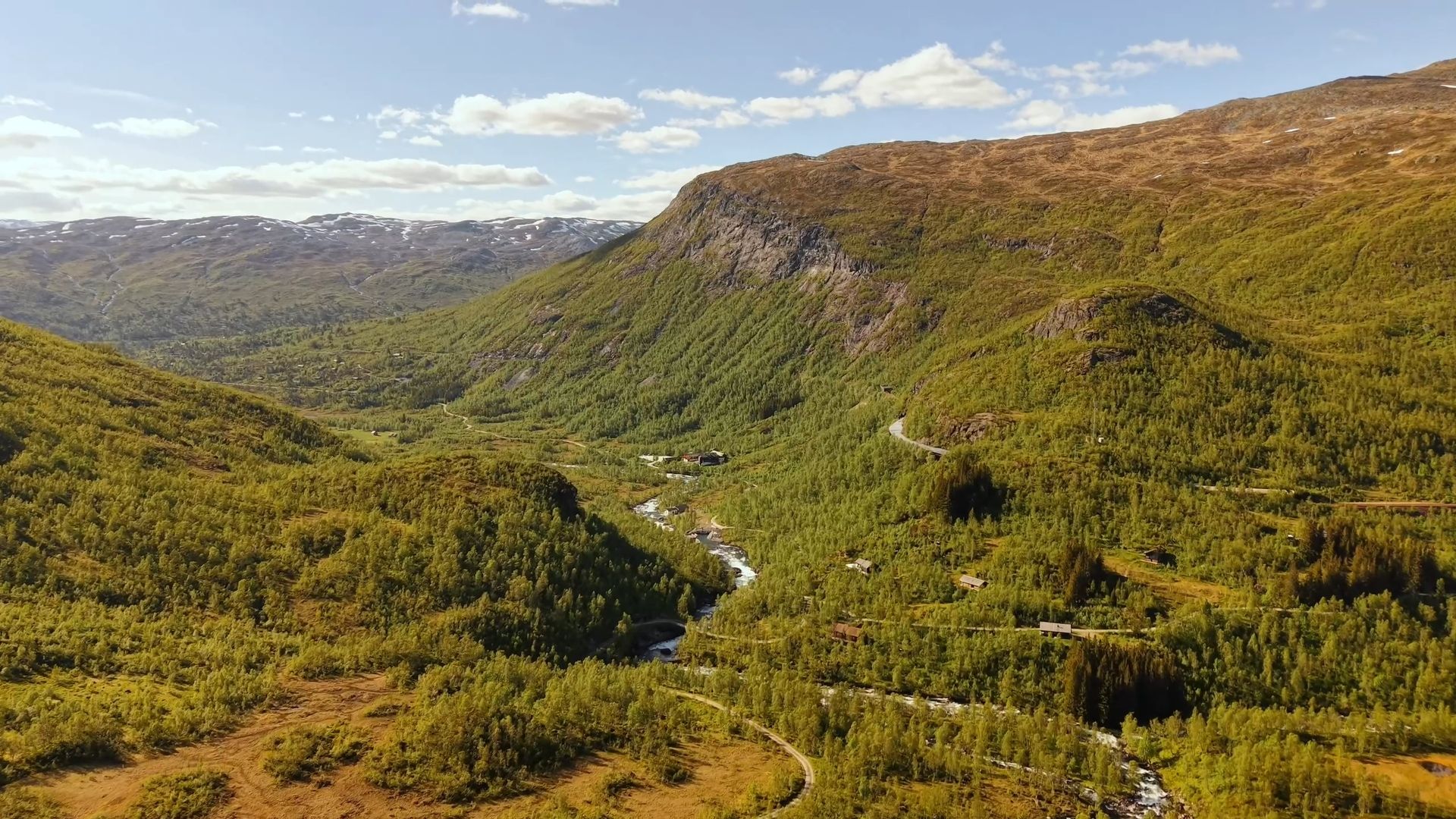 Mountain valley with a river, forested slopes, winding roads, and distant snow-dusted peaks.